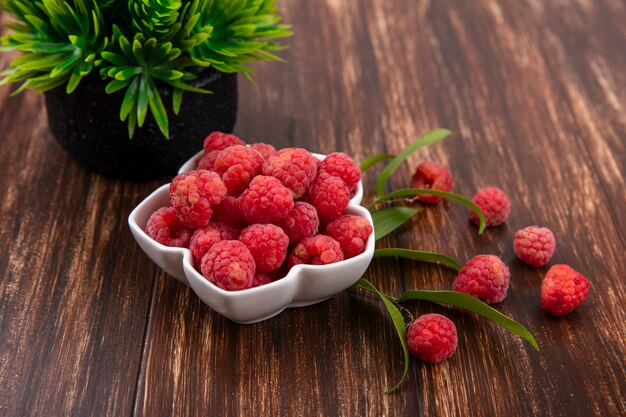 Side view of bowl of raspberry with flower and leaves on wooden surface