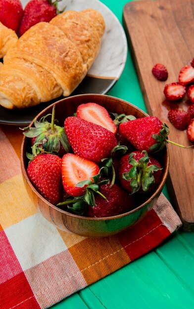Side view of bowl of strawberries with crescent roll in plate on cloth on green table