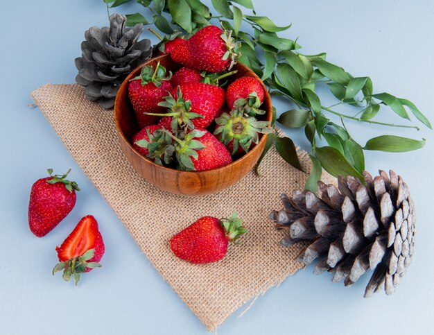 Side view of bowl of strawberries with pinecones on sackcloth on white table decorated with leaves