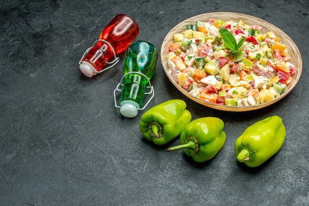 Vista laterale della ciotola di insalata di verdure con peperoni e bottiglie di olio e aceto sul tavolo verde scuro