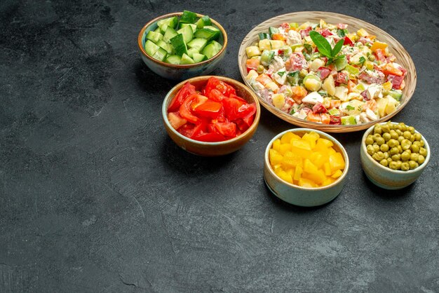 Side view of bowl of vegetable salad with bowls of vegetables on side on dark green background