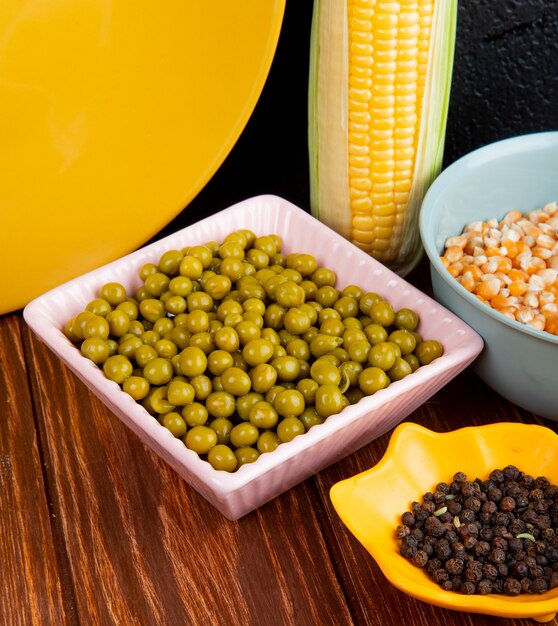 Side view of bowls of green peas corn seeds and black pepper on wooden table