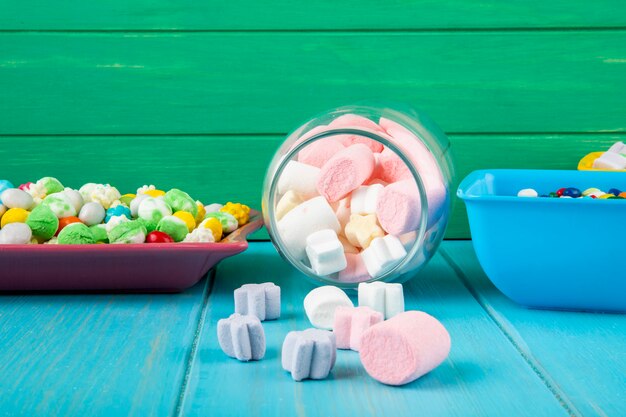 Side view of bowls with various colorful candies and marshmallow scattered from a glass jar on blue background