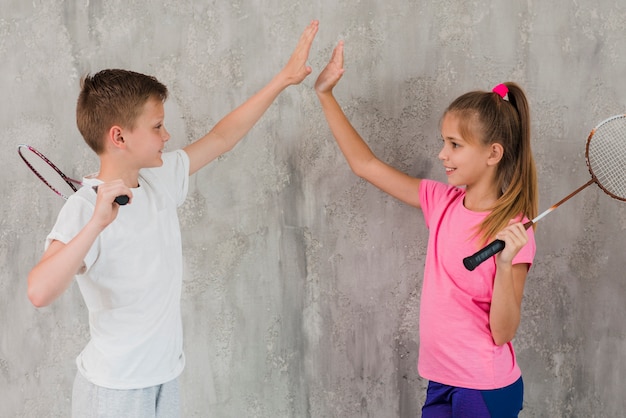Side view of a boy and girl holding racket in hand giving high five standing against wall