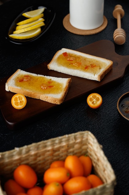 Side view of bread slices with jam smeared on them on cutting board with lemon slices kumquats in basket with clotted milk and honey dipper on black background