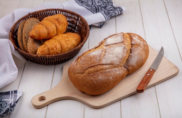 布のバスケットにクロワッサンと種をまく茶色の穂軸のパンのスライスとしてのパンと木製の背景のまな板にナイフで無愛想なパンの側面図