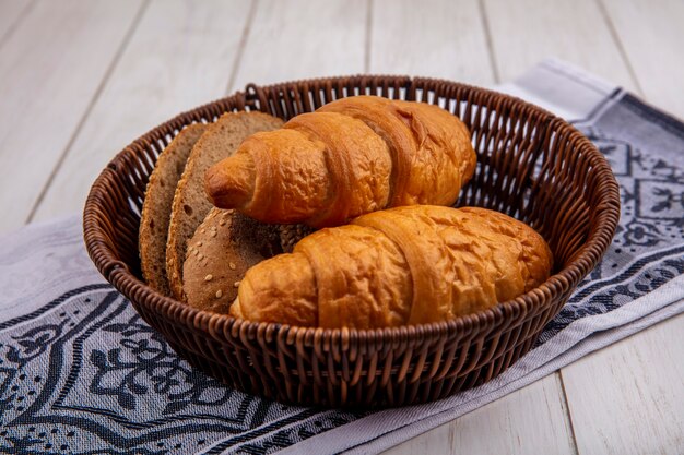 Side view of breads as croissant and seeded brown cob bread slices in basket on cloth on wooden background