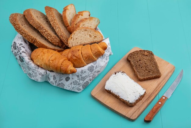 Side view of breads as sliced seeded cob baguette and croissant in bowl and rye bread smeared with cheese on cutting board with knife on blue background