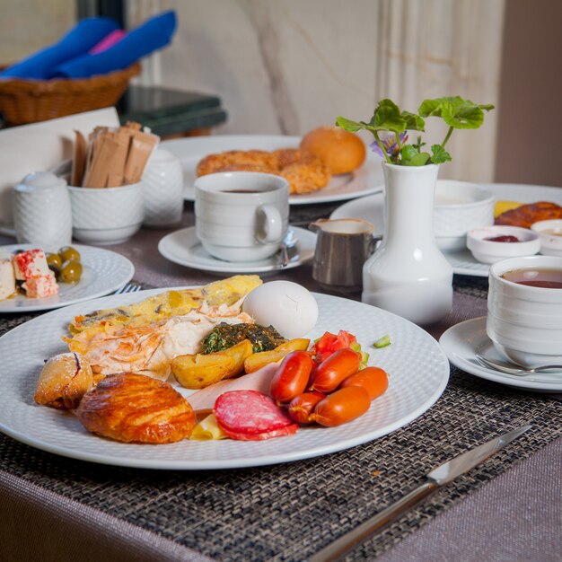 サイドビューの朝食ソーセージ、ゆで卵、オムレツ、プレートのクロワッサン、テーブルの上のお茶のカップ