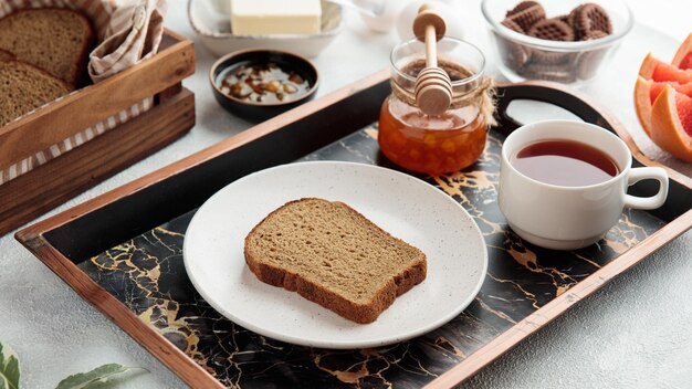 Вид сбоку на набор для завтрака с ломтиком хлеба, джемом из айвы, чашкой чая в подносе с печеньем, грейпфрутовым маслом на белом фоне