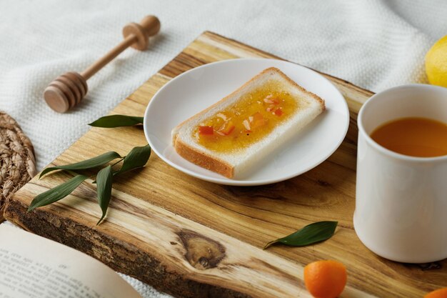 Side view of breakfast set with bread slice smeared with jam in plate and cup of orange juice kumquat slice with leaves on cutting board and jam dipper trivet and open book on white cloth background