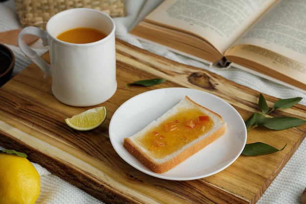 Side view of breakfast set with bread slice smeared with jam in plate and cup of orange juice lime slice with leaves on cutting board and lemon with open book on white cloth background