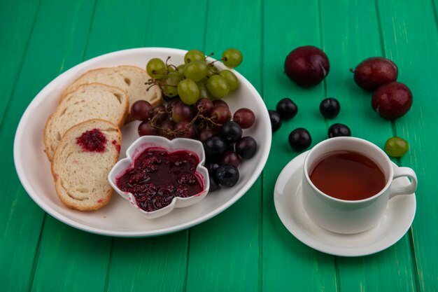 Вид сбоку на набор для завтрака с ломтиками хлеба, малиновым джемом и виноградом в тарелке и чашкой чая с плуотами на зеленом фоне
