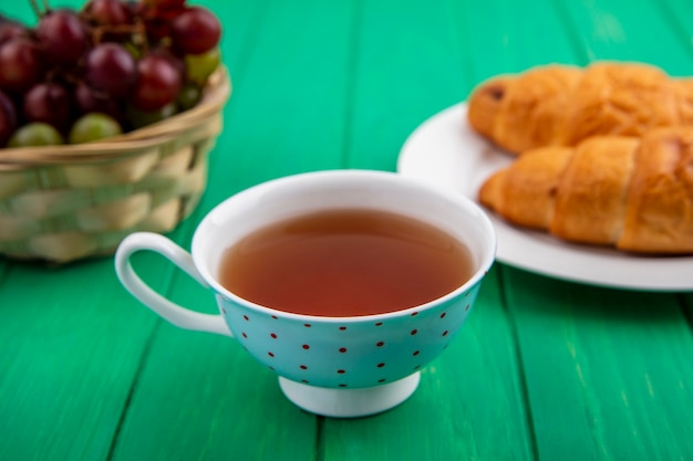Вид сбоку на набор для завтрака с круассанами в тарелке с чаем и корзиной ягод виноградного терна на зеленом фоне