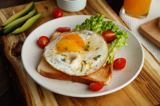 Side view of breakfast set with fried egg lettuce tomatoes on dried bread slice in plate and cucumber slices on cutting board with orange juice on cloth on wooden background
