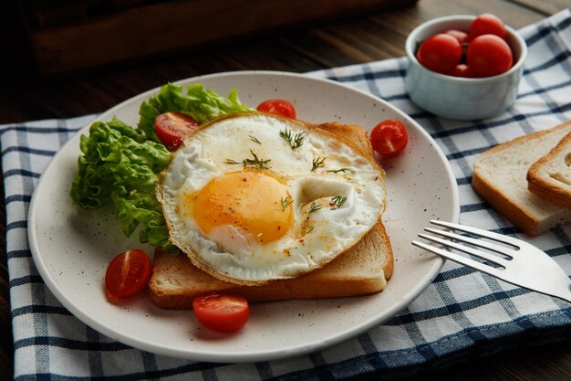 Side view of breakfast set with fried egg lettuce tomatoes on dried bread slice in plate with fork on plaid cloth on wooden background