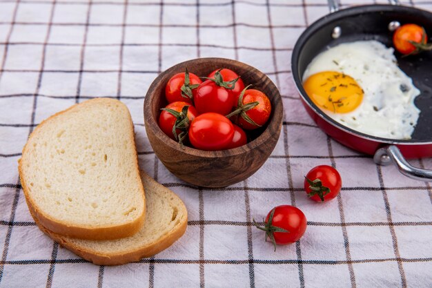 チェック柄の布の表面に目玉焼きのパンとパンのスライスとトマトのボウルで朝食セットの側面図