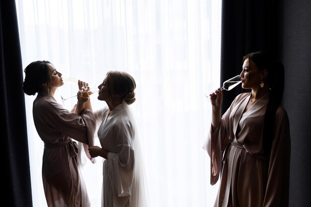 Вид сбоку невесты и подружки невесты с макияжем и прической в бело-розовой ночной рубашке, стоящих у окна и имеющих