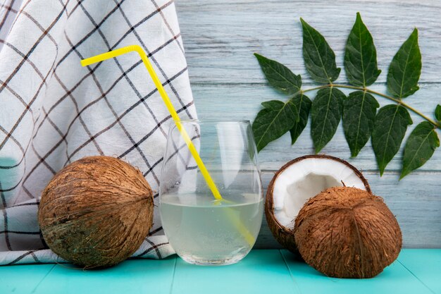 Side view of brown coconuts with a glass of water and leaf on tablecloth and grey surface