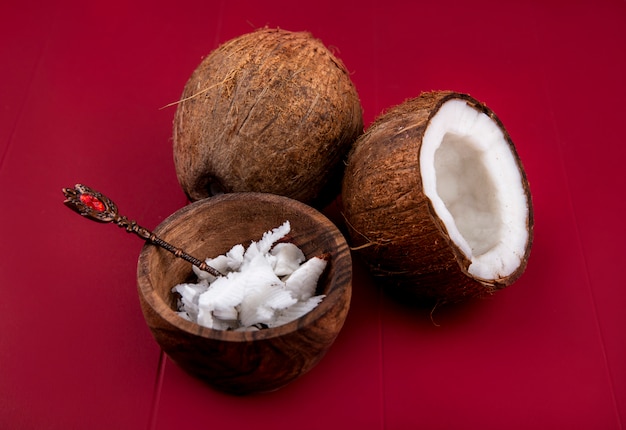 Side view of brown coconuts with whole and halved coconut pulps of coconut in a wooden bowl on red surface