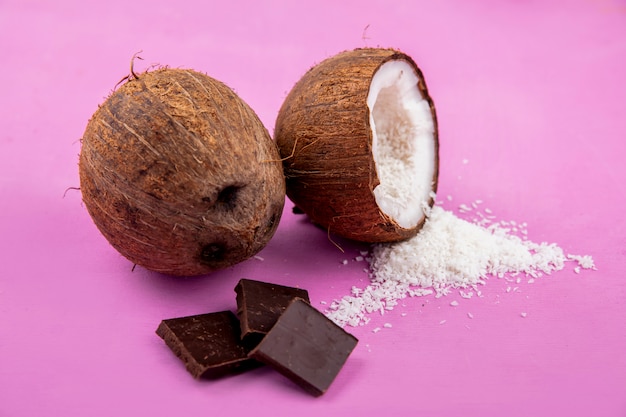 Вид сбоку коричневых и свежих кокосов с кокосовым порошком и шоколадной плиткой на розовой поверхности