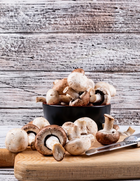 Side view brown and white mushrooms in bowl and on cutting board with knife on light wooden table