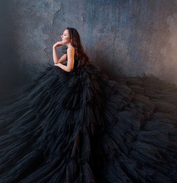 Side view of brunette in a lush black dress sitting on a background of the wall