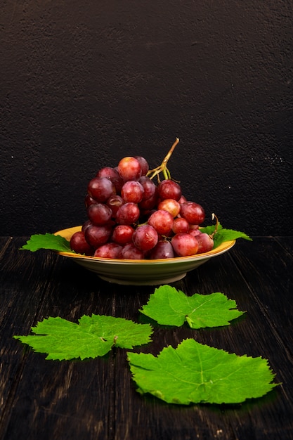 Side view of a bunch of sweet grapes in a plate and green grape leaves on rustic table