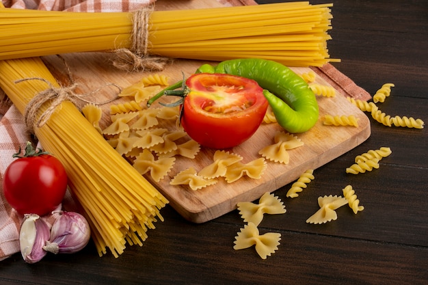 Side view of bunches of raw pasta with garlic and pasta with tomatoes and hot pepper on a board on a wooden surface