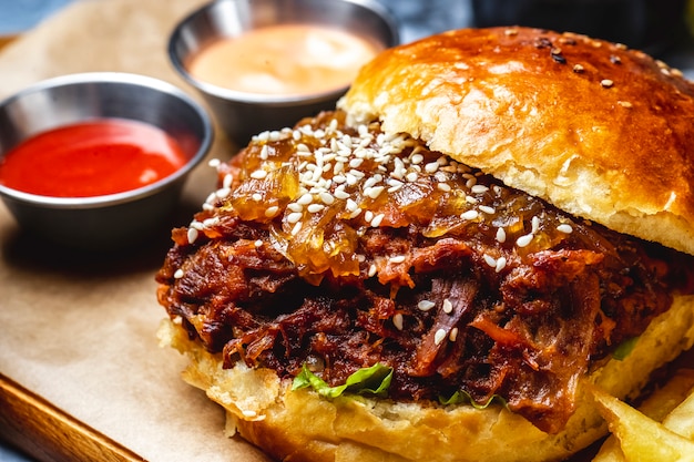 Side view burger with minced meat caramelized onion sesame seeds lettuce between burger bun and sauces on the table