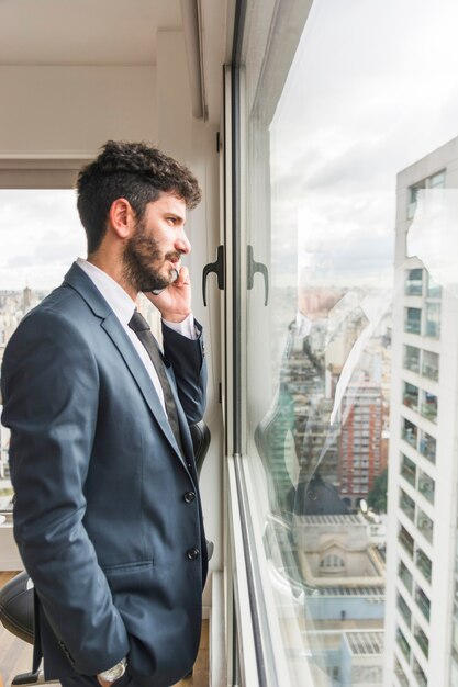 Vista laterale dell'uomo d'affari che sta vicino alla finestra dell'ufficio che parla sul telefono cellulare