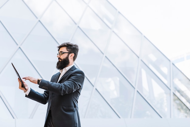 Вид сбоку бизнесмена с длинной бородой, используя цифровую табличку, стоящую против стеклянного здания