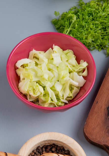 Side view of cabbage slices in bowl with bunch of coriander and black pepper seeds on blue background