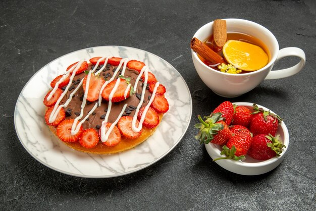 Side view cake with a cup of tea cake with strawberries and chocolate next to the bowl of strawberries and the cup of tea with lemon and cinnamon sticks on the black table