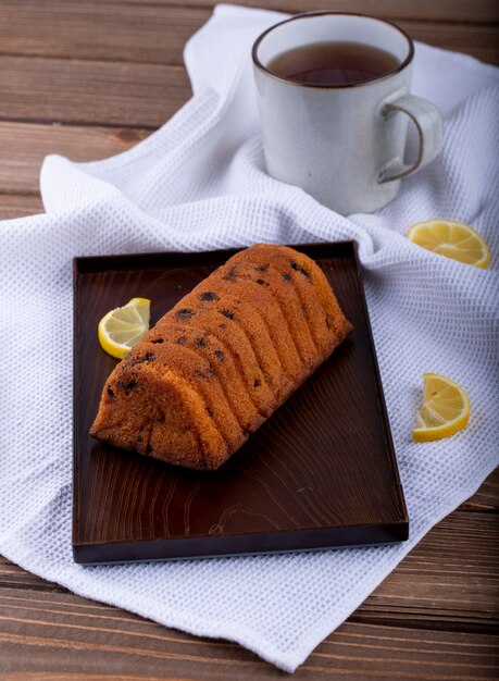 Side view of cake with raisins and lemon slices on a wooden board and a mug of tea on the tablecloth