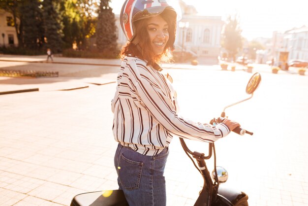 Side view of carefree african woman rides on modern motorbike outdoors and looking at the camera