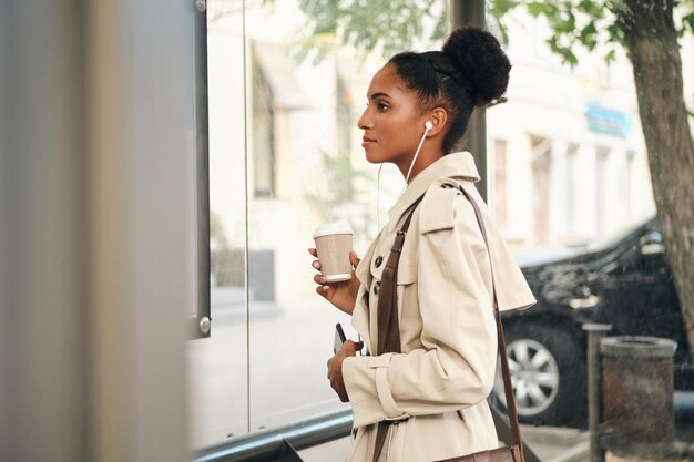 Side view of casual African American girl in stylish trench coat with coffee to go and cellphone watching route at bus stop