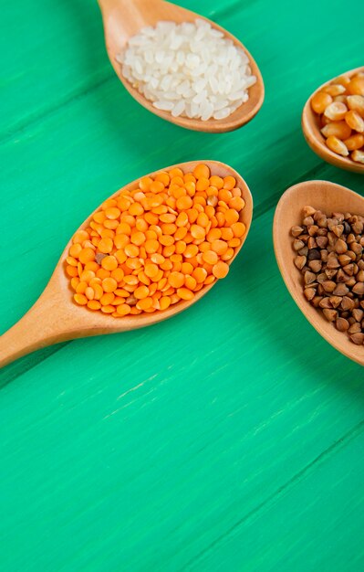 Side view of cereal grains and seeds in wooden spoons corns rice buckwheat and red lentils on green table