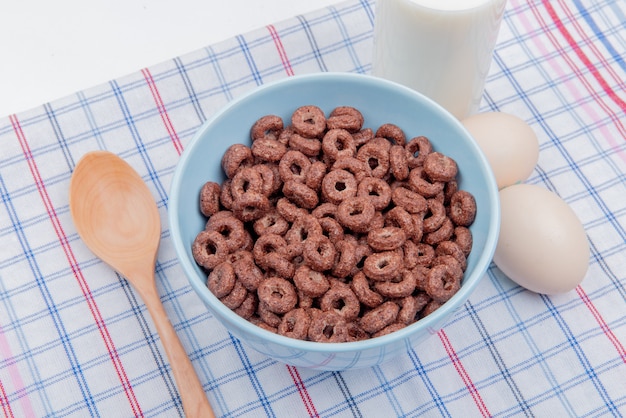 Free photo side view of cereals in bowl with milk eggs and wooden spoon on plaid cloth and white