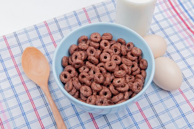 side view of cereals in bowl with milk eggs and wooden spoon on plaid cloth and white