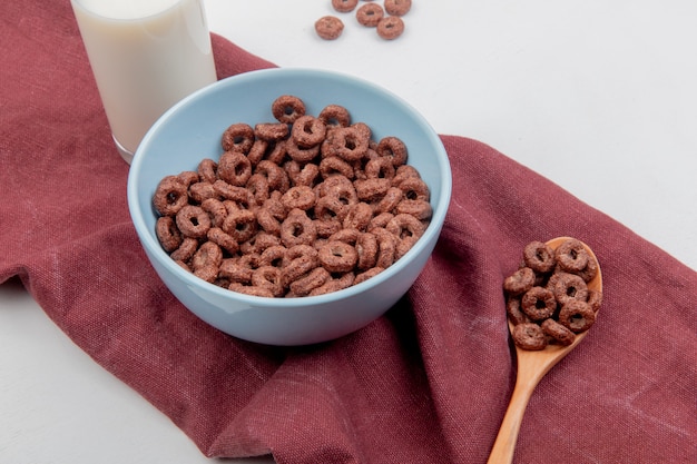 Side view of cereals in bowl and in wooden spoon with milk on bordo cloth and white surface