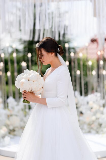 Вид сбоку очаровательной невесты брюнетки в вуали и элегантном свадебном платье, смотрящей на букет пионов, стоящих на цветущем алтаре Свадебная церемония на открытом воздухе Красивая невеста
