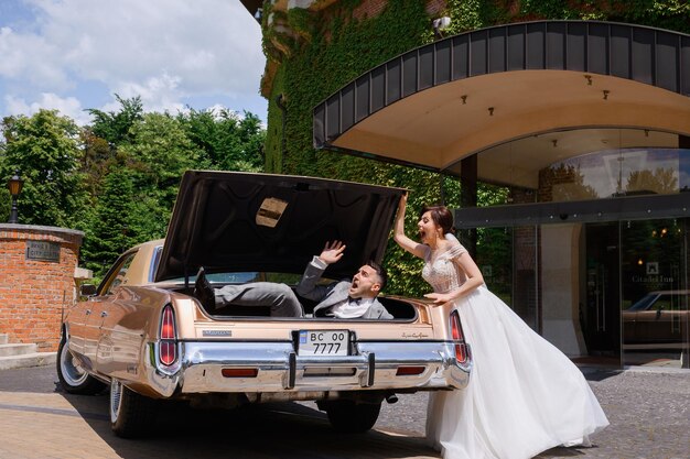 Side view of cheerful bride in beautiful wedding dress standing opposite retro car on hotel area feeling fun while closing