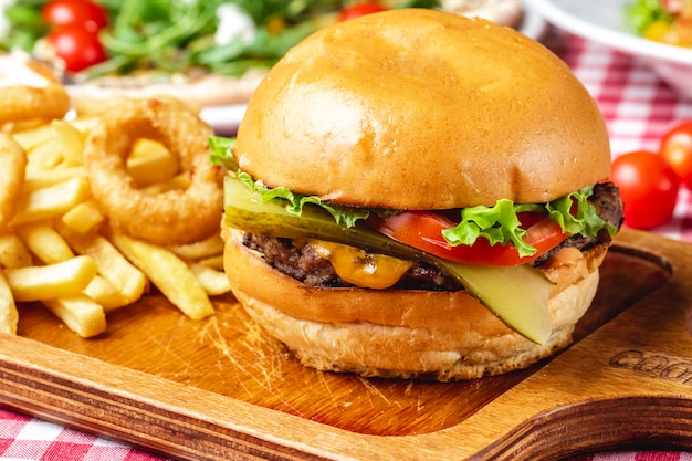 Side view cheeseburger grilled beef patty pickled cucumber fresh tomato lettuce cheese between burger buns french fries and onion rings on the table