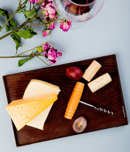 Side view of cheeses as cheddar and parmesan with grape corks and corkscrew on cutting board and flowers on white 1