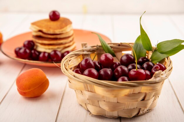 Side view of cherries in basket and plate of pancakes and cherries with apricot on wooden background