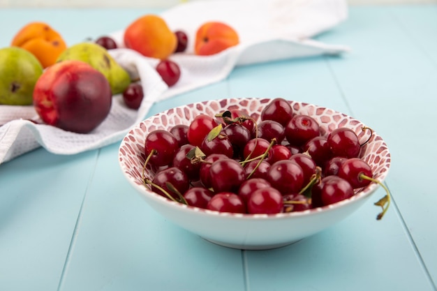 Side view of cherries in bowl and pattern of peach pear apricot cherry on white cloth on blue background