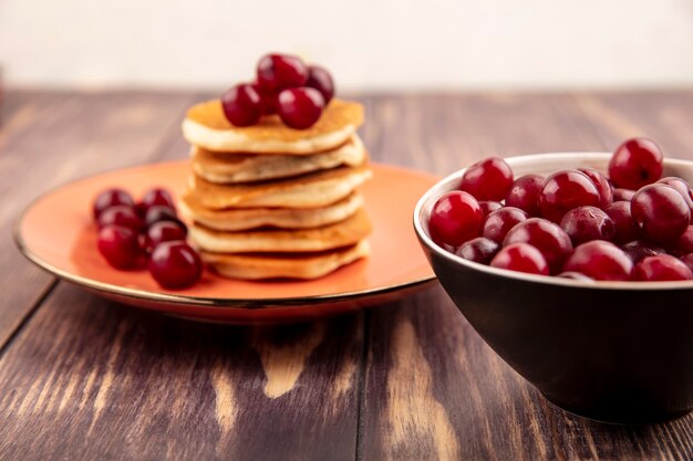 Side view of cherries in bowl and plate of pancakes and cherries on wooden surface and white background