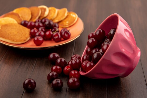 Side view of cherries spilling out of bowl and plate of pancakes and cherries on wooden background