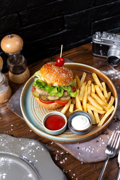 Side view of chicken burger with pickles and tomatoes served with french fries and sauces on dark